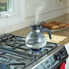 Clear borosilicate glass whistling teakettle with black handle and stainless steel band boiling on a gas stove, ideal for Kitchen & Food, Brewing Coffee and Tea, Best Sellers, and Promo Eligible collections.