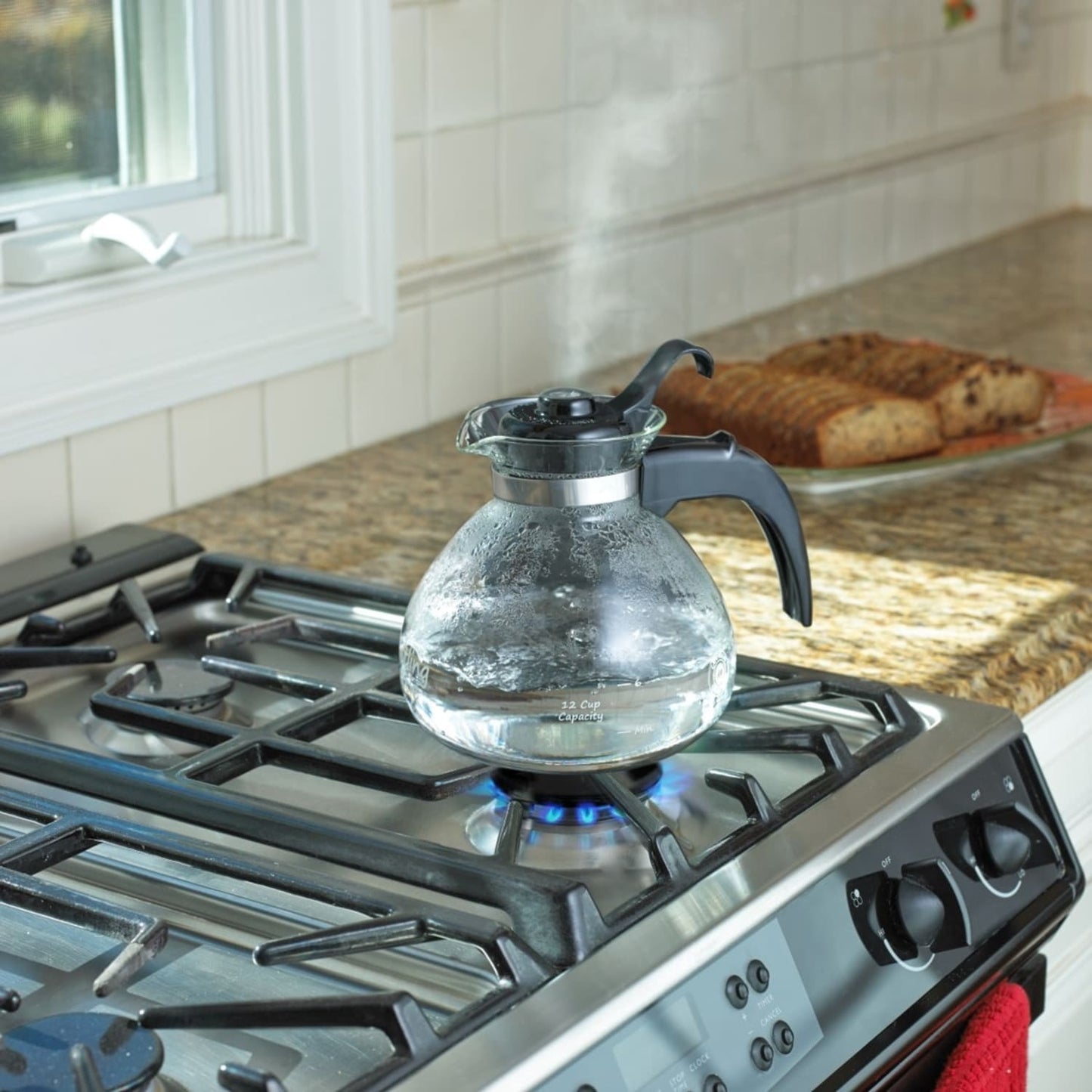 Clear borosilicate glass whistling teakettle with black handle and stainless steel band boiling on a gas stove, ideal for Kitchen & Food, Brewing Coffee and Tea, Best Sellers, and Promo Eligible collections.