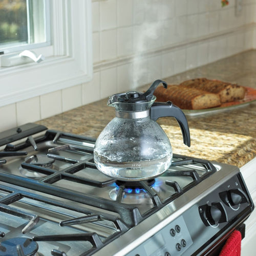 Clear borosilicate glass whistling teakettle with black handle and stainless steel band boiling on a gas stove, ideal for Kitchen & Food, Brewing Coffee and Tea, Best Sellers, and Promo Eligible collections.