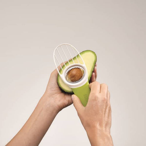 Person using a green 3-in-1 tool with plastic and metal parts to remove the pit from a halved avocado, ideal for food prep in Kitchen & Food and Farm, Garden & Tool collections.