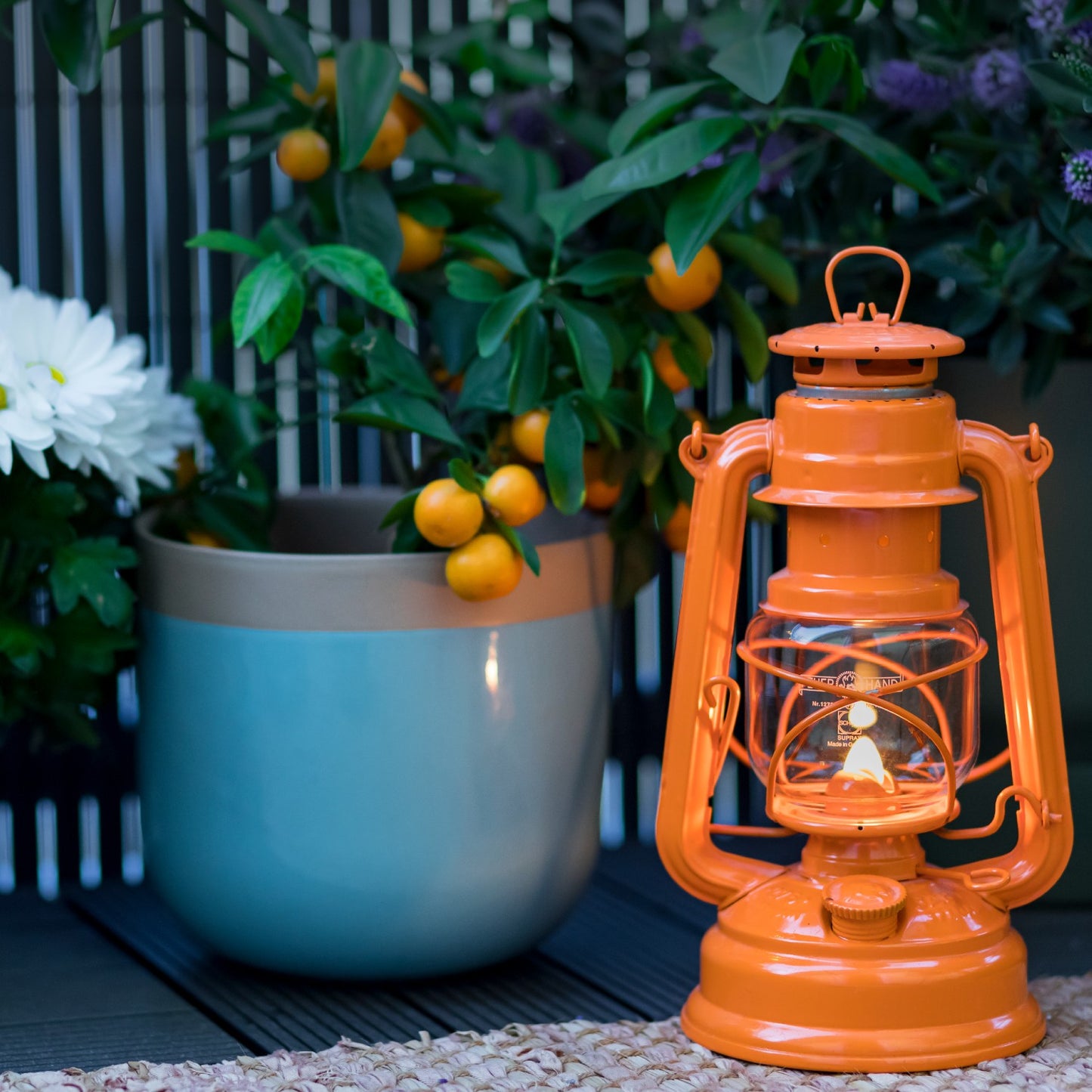 Handcrafted bright orange galvanized oil lantern glowing softly on outdoor deck next to potted small orange fruit tree, ideal for Lamps & Lighting, Camping, Emergency Supplies collections.