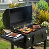 Outdoor charcoal grill with an open lid showing a seasoned steel grilling pan filled with colorful vegetables and adjacent steaks, surrounded by greenery in a backyard setting.