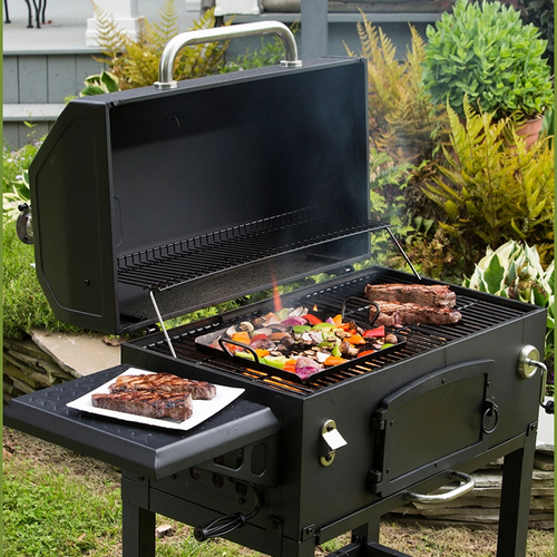 Outdoor charcoal grill with an open lid showing a seasoned steel grilling pan filled with colorful vegetables and adjacent steaks, surrounded by greenery in a backyard setting.