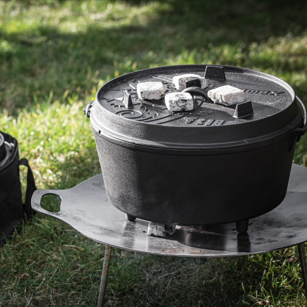 Robust cast iron Dutch oven with three legs heating outdoors on a metal stand, featuring a fitted embossed lid with charcoal briquettes for even cooking, perfect for Kitchen & Food, Cookware, Cast Iron, Best Sellers, Promo Eligible collections.