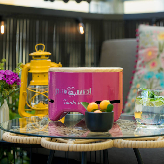 Handcrafted pink tabletop charcoal grill with wooden lid, set on a glass and woven outdoor table alongside a yellow lantern, small bowl of fruit, and glass of iced beverage, perfect for Farm, Garden & Tool, Camping, and Grilling collections.