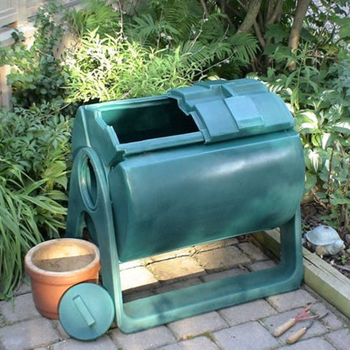 Green plastic compost tumbler with hinged lid open, positioned on a paved garden area surrounded by plants and gardening tools, suitable for outdoor composting in Appliances and Composting Toilets collections.