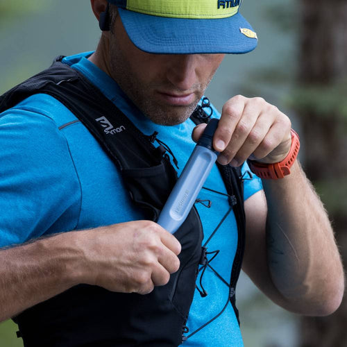 Man wearing a blue shirt and hat outdoors using a handheld water filter straw from his backpack, part of camping and emergency supplies for clean water access.
