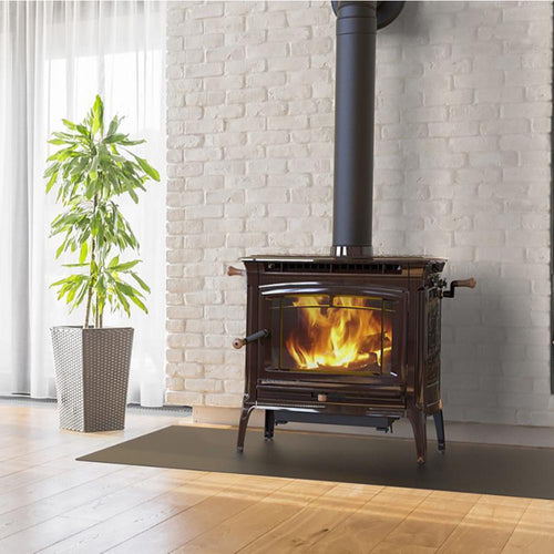 Dark brown wood-burning stove with cast iron legs and stainless steel handles, featuring a visible roaring fire inside, set against a white brick wall on a protective mat, in a bright room with wooden floors and a green potted plant nearby, ideal for Stoves, Heatstoves, and Hearthstone Wood Heatstoves collections.