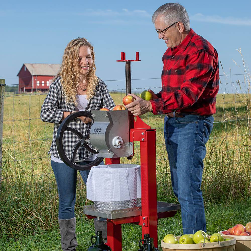 Man and woman using a handcrafted stainless steel cider press with red frame and black crank wheel in a farm setting, featured in Farm, Garden & Tool and Apple Harvest Supplies collections.