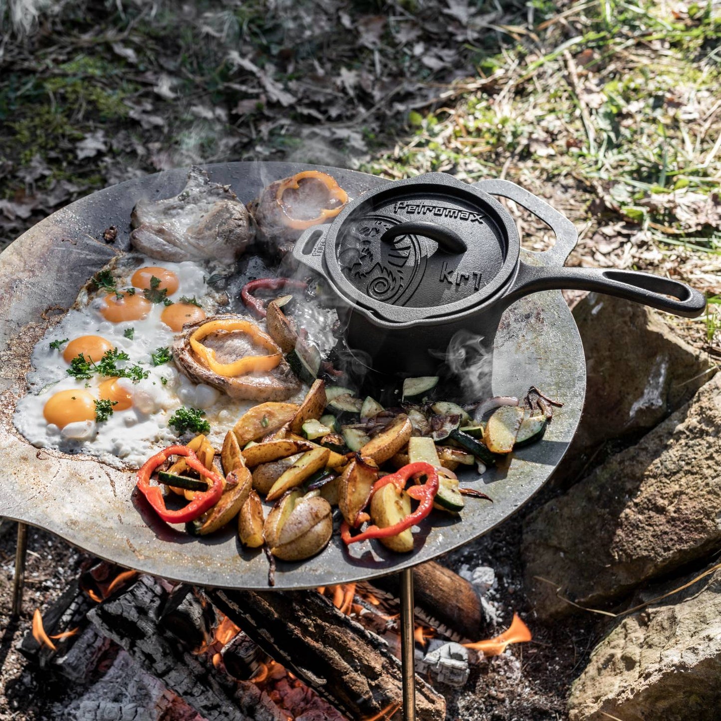 Handcrafted cast iron saucepan with lid cooking over an open fire alongside roasted potatoes, bell peppers, mushrooms, and eggs, ideal for camping and outdoor kitchen use.