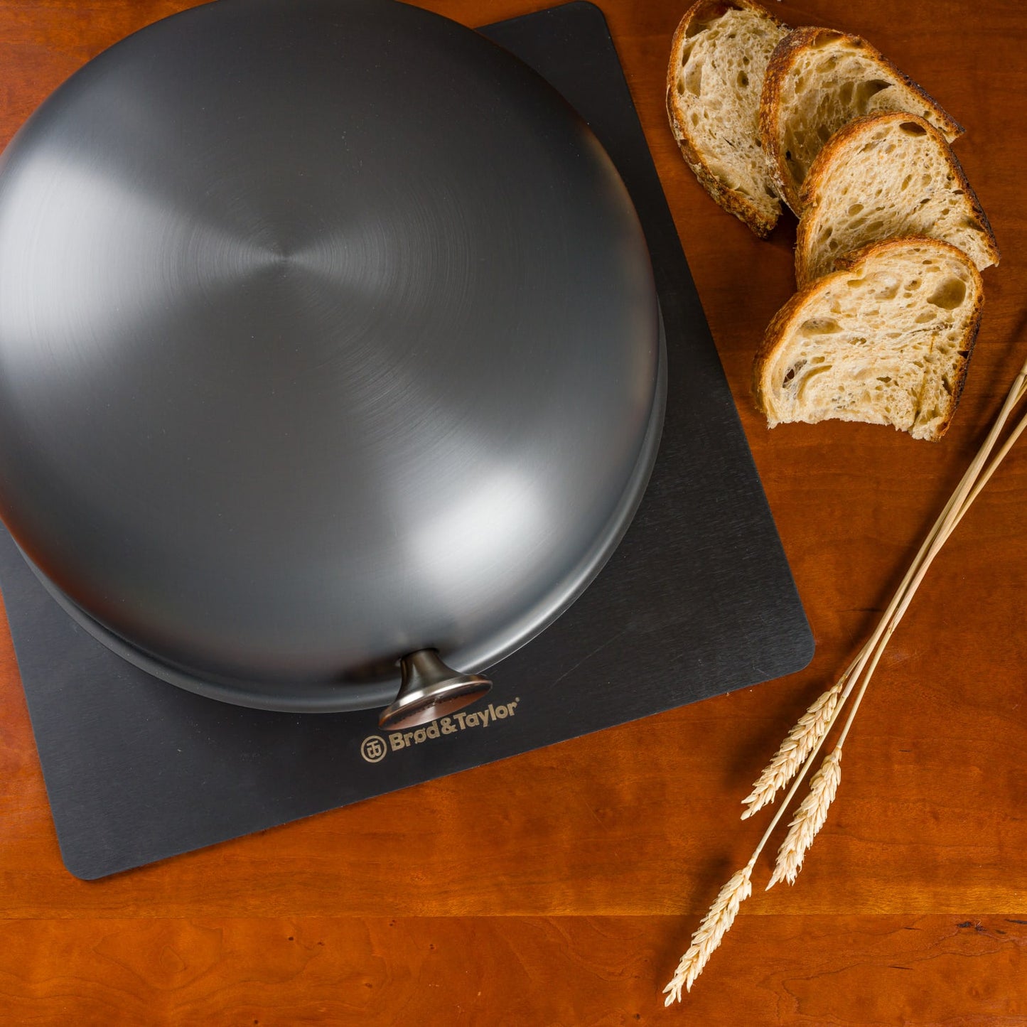Matte black Brod & Taylor Baking Shell with smooth curved surface placed on a branded black mat next to sliced artisan bread and wheat stalks, suitable for Kitchen & Food and Baking Supplies collections.