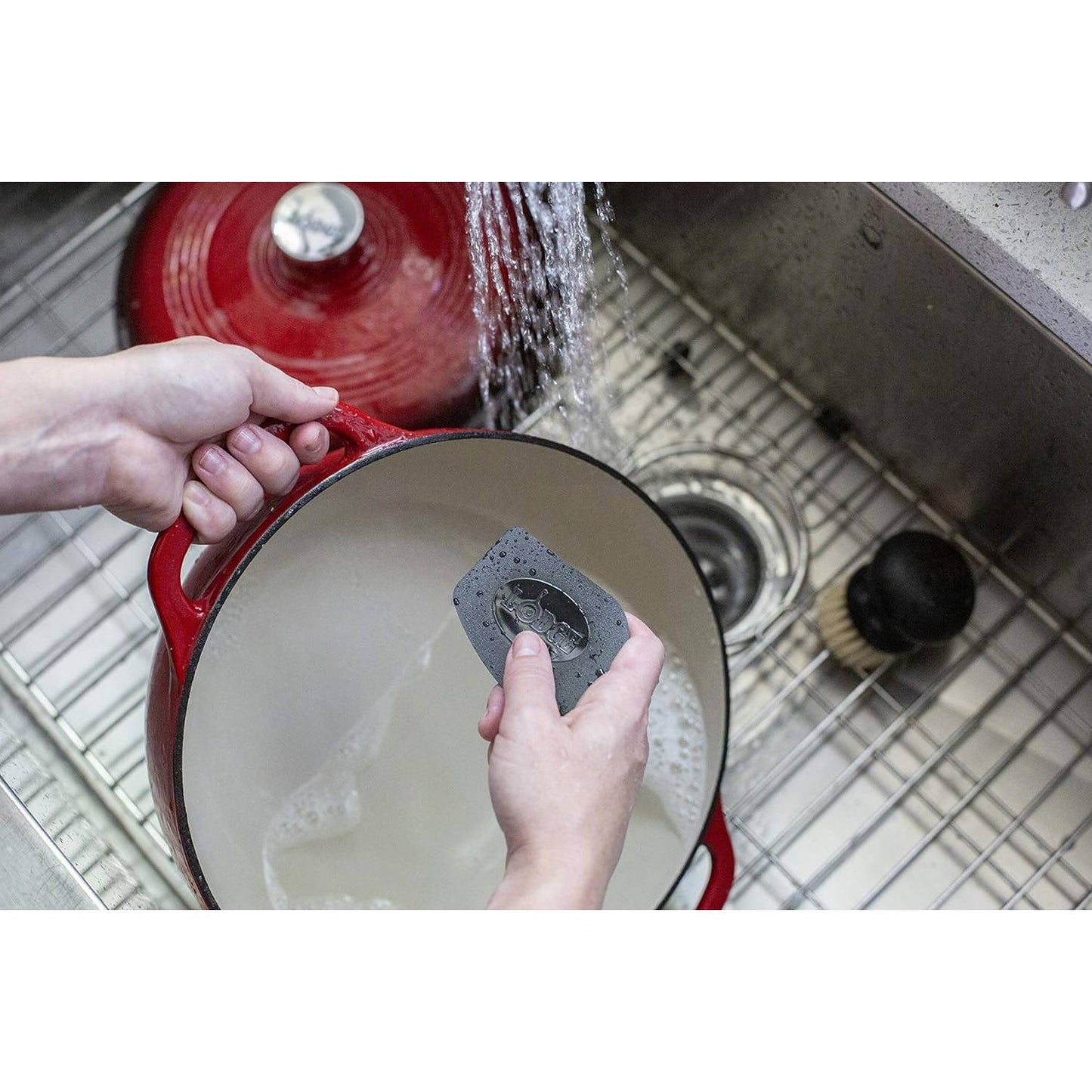 Hands washing red enameled cast iron pot in a kitchen sink using a polycarbonate pan scraper under running water, showcasing essential care tools for cast iron cookware in Kitchen & Food, Cookware collections.