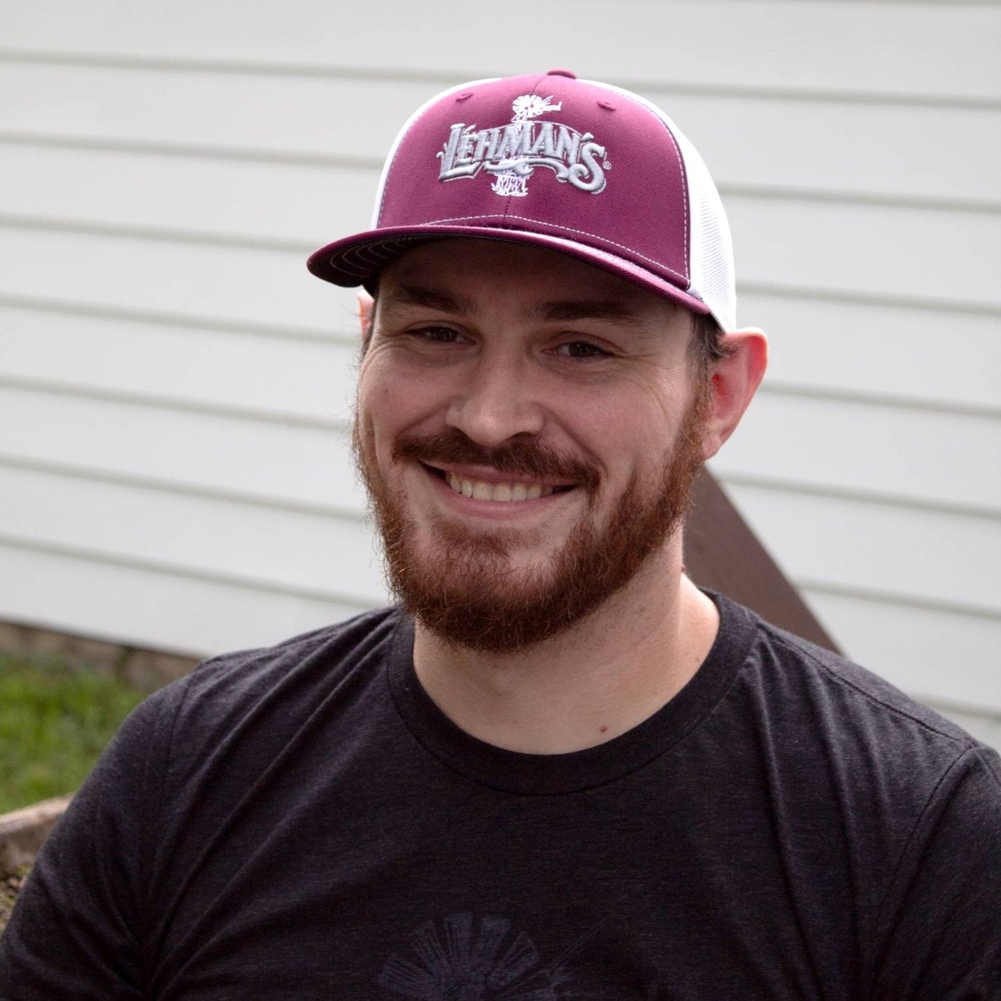Man smiling outdoors wearing a handcrafted Lehman's embroidered trucker hat with a burgundy front panel and white stretch mesh back, featured in Health & Wellness, Clothing and Accessories, Best Sellers, and Promo Eligible collections.