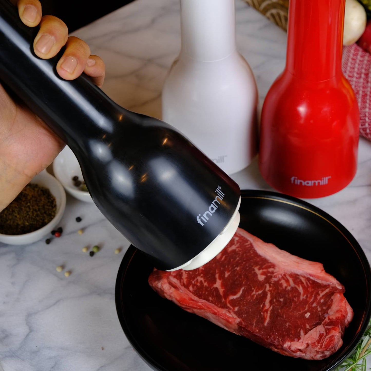 Handheld black pepper grinder actively seasoning a raw steak on a plate, with additional white and red grinders nearby, ideal for Kitchen & Food and Kitchen Gadgets collections.