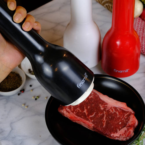 Handheld black pepper grinder actively seasoning a raw steak on a plate, with additional white and red grinders nearby, ideal for Kitchen & Food and Kitchen Gadgets collections.
