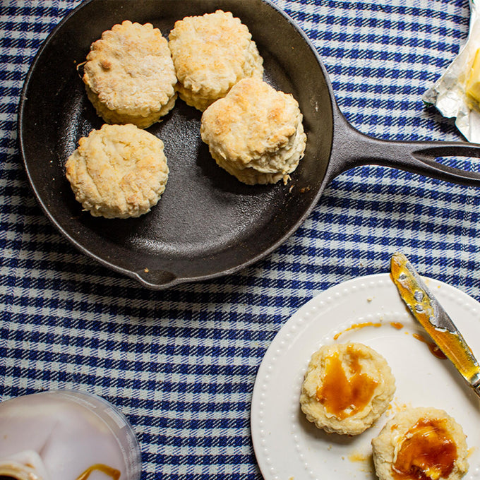 Handcrafted Lodge Classic Cast Iron Skillet with four golden biscuits cooking on a blue checkered cloth, accompanied by a plate with buttered and syrup-topped biscuits, ideal for Kitchen & Food, Cookware, Cast Iron, Best Sellers, and Promo Eligible collections.