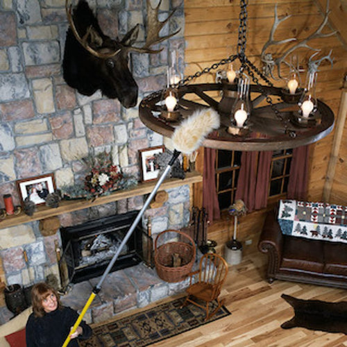 Person using a Lamb's Wool Angled Telescoping Duster with a long handle to clean a rustic wagon wheel chandelier in a cozy lodge-style living room, featuring stone fireplace, wooden walls, and decorative antlers, part of Home & Decor, Household Cleaning, Cleaning Utensils and Gadgets, Best Sellers, and Promo Eligible collections.
