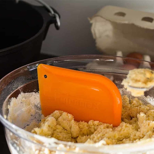 Orange pan scraper partially embedded in dough inside a glass bowl, showcasing kitchen tool use for baking in Kitchen & Food and Cookware collections.