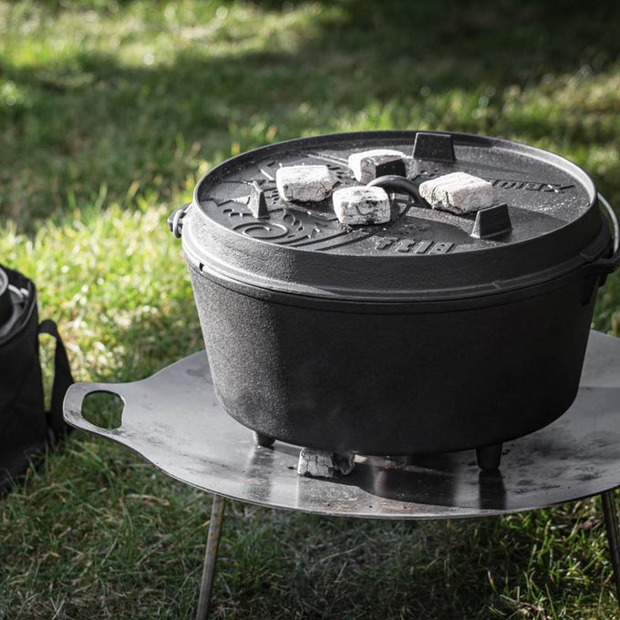 Heavy-duty cast iron pot with legs and a fitted lid holding charcoal briquettes on top, perfect for outdoor cooking and baking, featured in Kitchen & Food, Cookware, and Cast Iron collections.