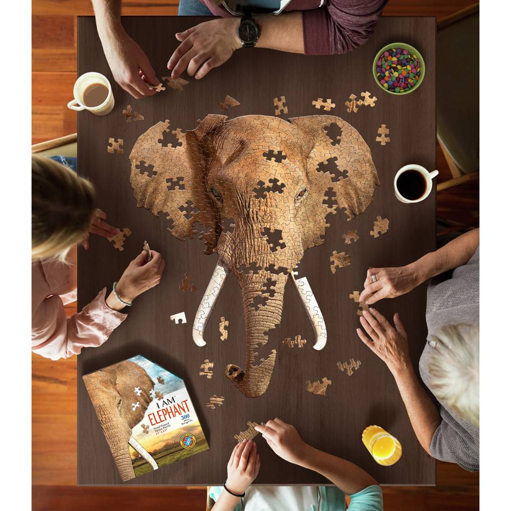 Group of four people assembling a 300-piece elephant-shaped jigsaw puzzle on a wooden table, with coffee, juice, colorful candy, and the puzzle box nearby in a family-friendly game setting.