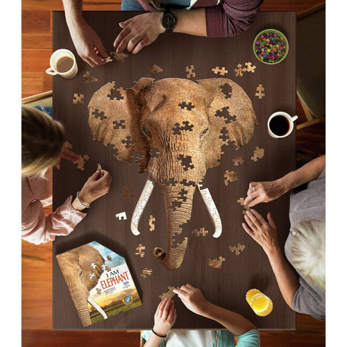 Group of four people assembling a 300-piece elephant-shaped jigsaw puzzle on a wooden table, with coffee, juice, colorful candy, and the puzzle box nearby in a family-friendly game setting.