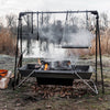 Portable steel fire trough suspended on a black metal frame with hanging grill grate over a wood fire, set outdoors near a lake in a natural Farm, Garden & Tool and Camping setting.