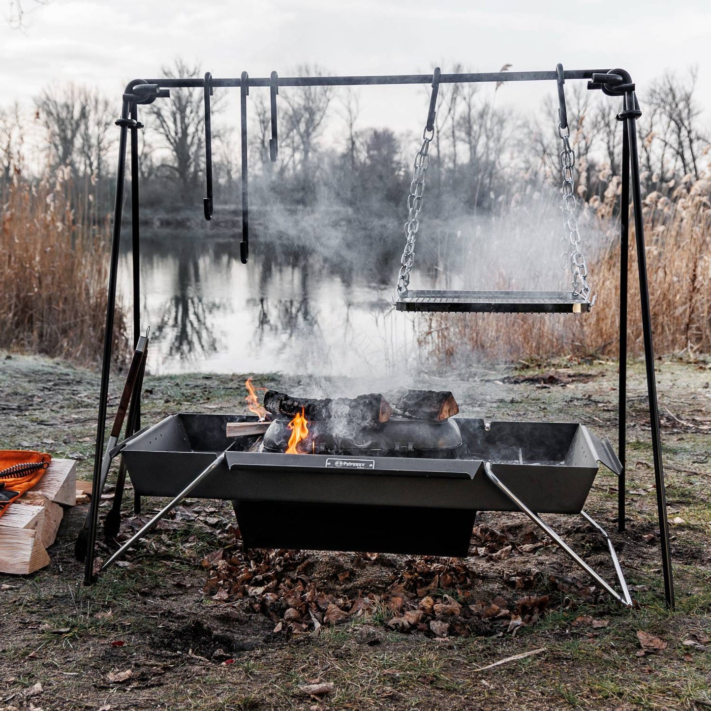 Portable steel fire trough suspended on a black metal frame with hanging grill grate over a wood fire, set outdoors near a lake in a natural Farm, Garden & Tool and Camping setting.