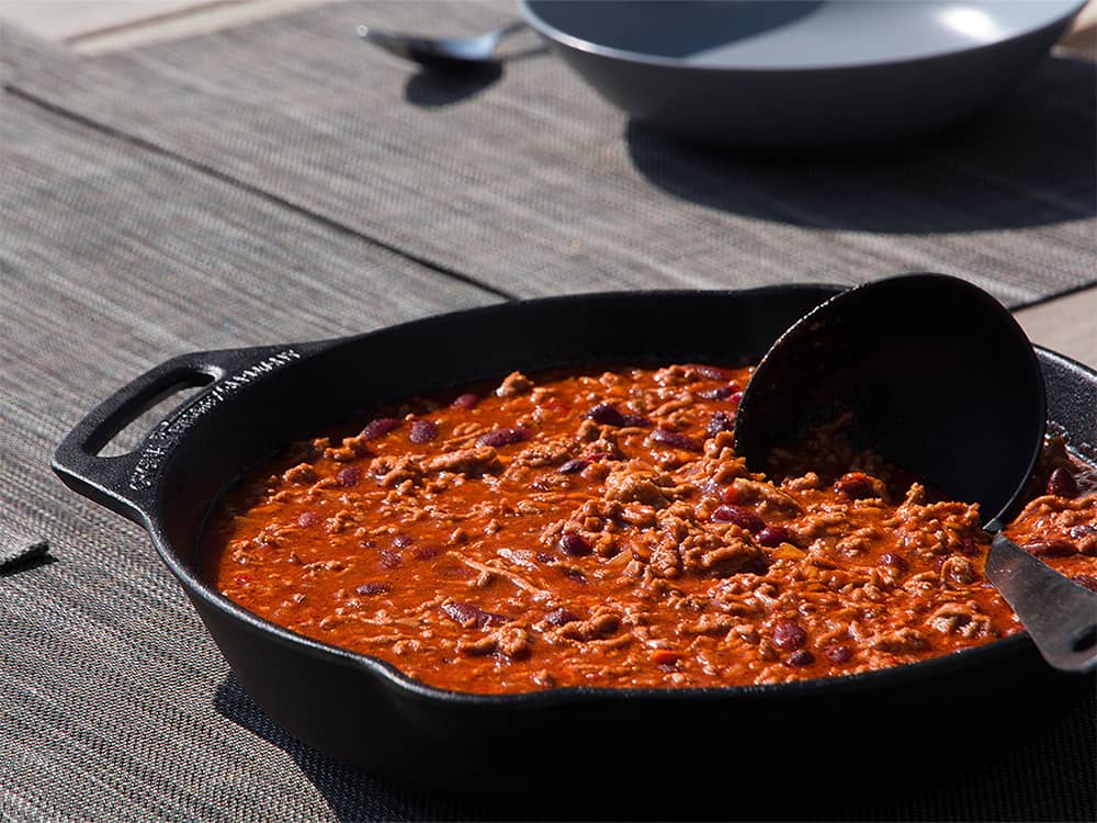 Cast iron skillet with a long handle and helper grip, filled with chili or stew, shown on an outdoor table, ideal for Farm, Garden & Tool, Camping, Kitchen & Food, and Cast Iron collections.