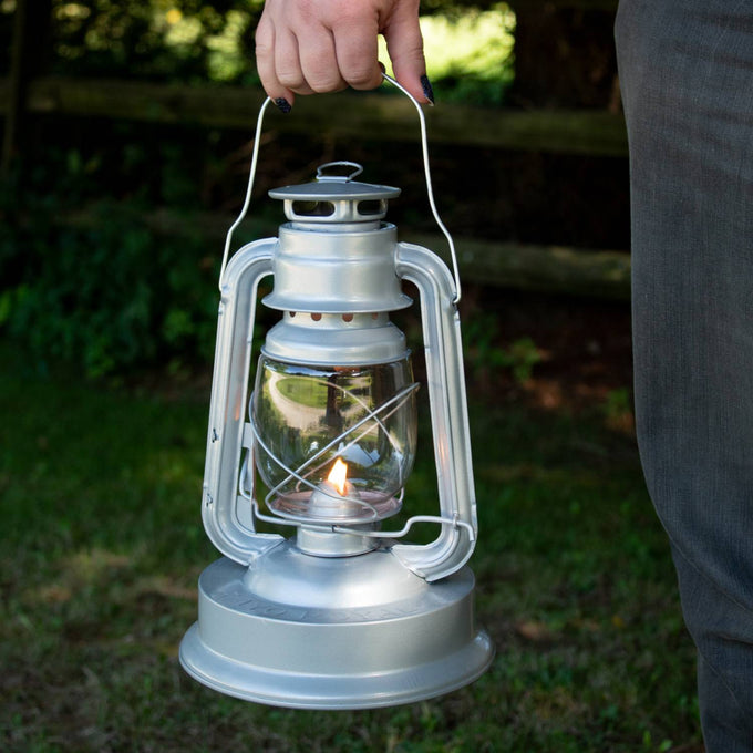 Handcrafted silver metal Rayo Royal Lantern with clear glass globe and protective wire frame, held outdoors, providing steady bright light for farm, garden, camping, and emergency use.