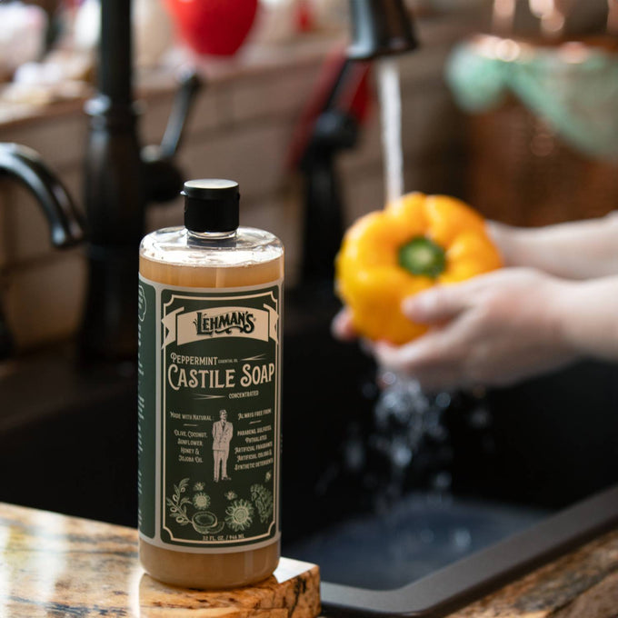 Clear bottle of Lehman's concentrated castile soap with peppermint scent next to a kitchen sink where a yellow bell pepper is being washed, highlighting handcrafted USA made soap in Health & Wellness, Grooming and Personal Hygiene, and Soaps collections.