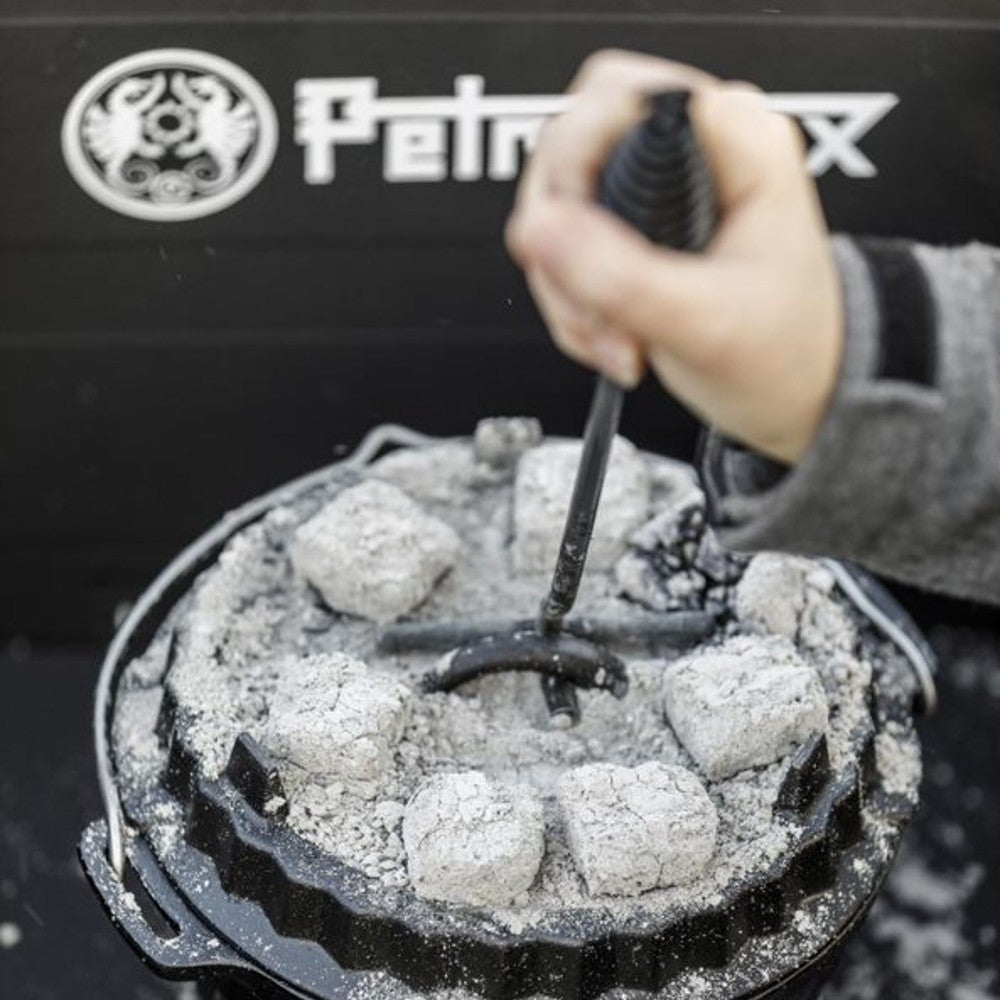 Hand holding a curved metal lid lifter with a coiled handle lifting a hot cast iron Dutch oven lid covered with glowing charcoal briquettes, featured in Farm, Garden & Tool, Camping, Best Sellers, and Promo Eligible collections.