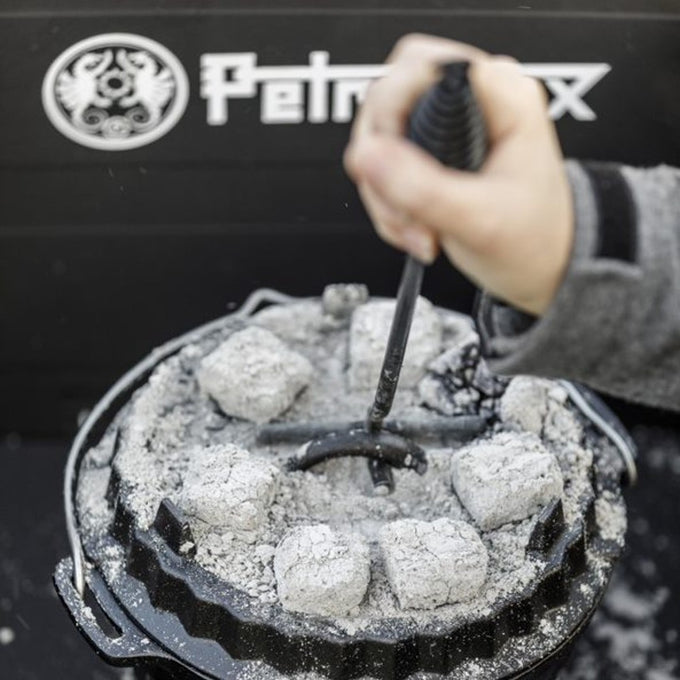 Hand holding a curved metal lid lifter with a coiled handle lifting a hot cast iron Dutch oven lid covered with glowing charcoal briquettes, featured in Farm, Garden & Tool, Camping, Best Sellers, and Promo Eligible collections.
