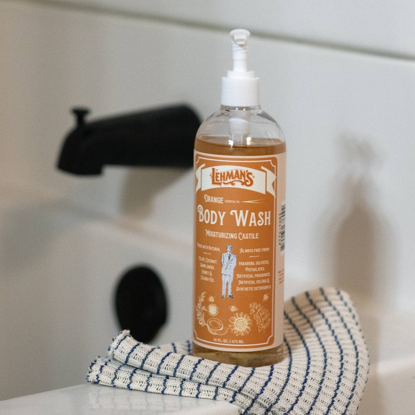 Clear plastic bottle of handcrafted moisturizing castile body wash with orange scent, featuring pump dispenser, placed on a striped cloth by a bathtub faucet, part of USA Made, Health & Wellness, Grooming and Personal Hygiene collections.