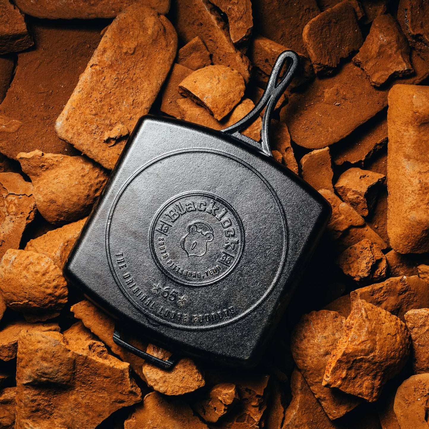 Square cast iron grill pan with embossed logo and triple-seasoned finish, displayed on rustic brown firewood pieces, highlighting handcrafted quality and USA-made cookware.