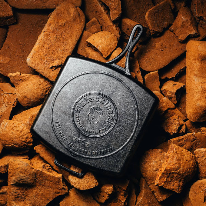 Square cast iron grill pan with embossed logo and triple-seasoned finish, displayed on rustic brown firewood pieces, highlighting handcrafted quality and USA-made cookware.
