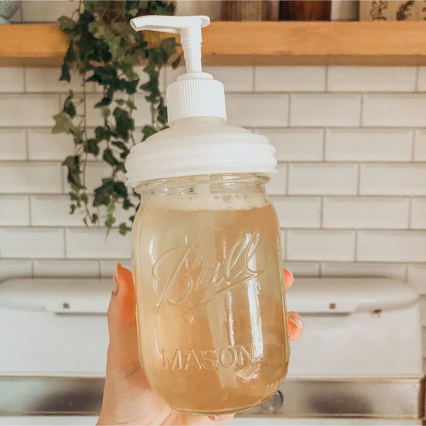 Hand holding a glass mason jar filled with soap and topped with a white pump lid, set against a farmhouse kitchen backdrop, highlighting eco-friendly Health & Wellness and Home & Decor collections.