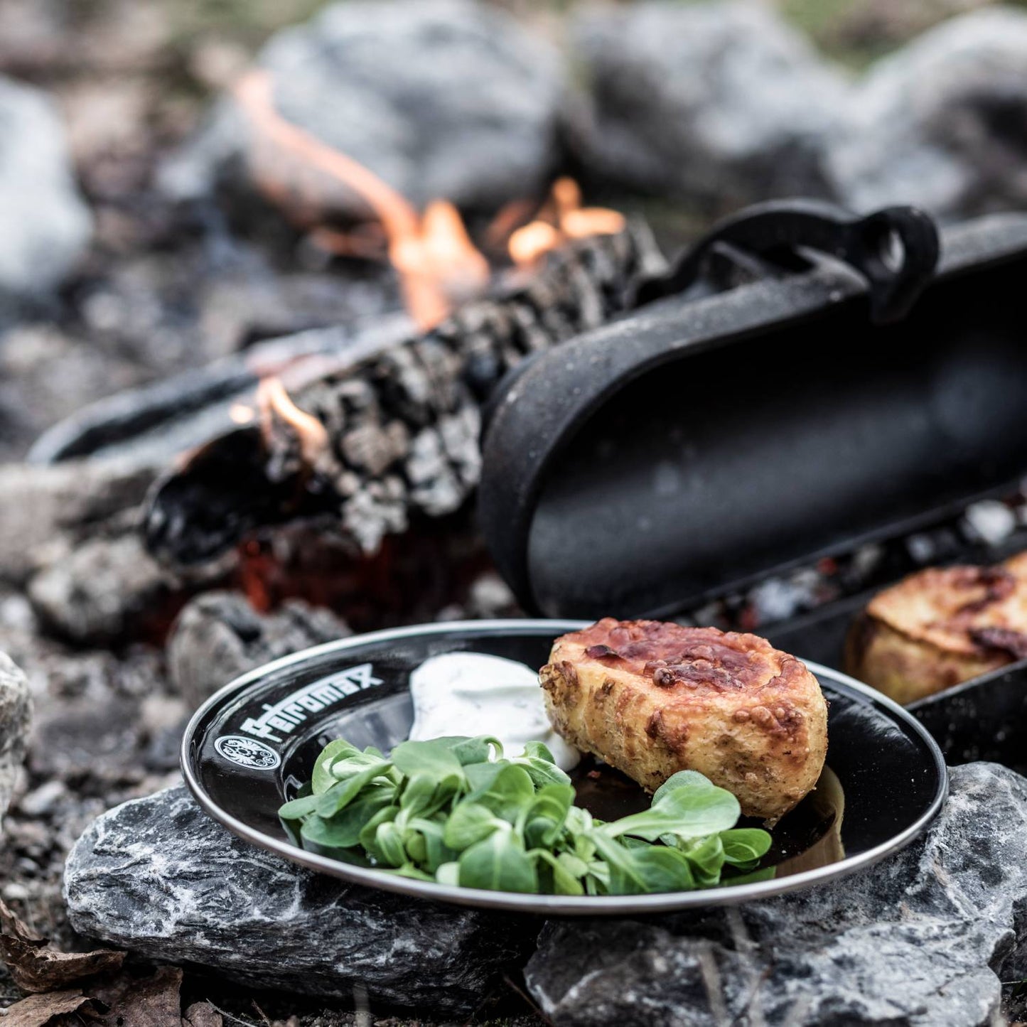 Black durable enamel plate holding fresh greens, a biscuit, and creamy sauce outdoors on rocks near a campfire, perfect for Farm, Garden & Tool, Kitchen & Food, Camping, Dinnerware, Best Sellers, and Promo Eligible collections.