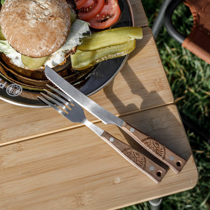 Serrated stainless steel knife and fork with engraved wooden handles resting on a bamboo table beside a sandwich plate, featured in Camping, Kitchen & Food, Farm, Garden & Tool collections.