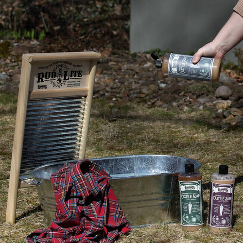 Handcrafted bottles of Lehman's Castile Concentrated Liquid Soap in an outdoor setting beside a metal wash tub, plaid cloth, and wooden washboard, highlighting USA made, natural soap for Health & Wellness, Grooming and Personal Hygiene, Soaps, and Best Sellers collections.