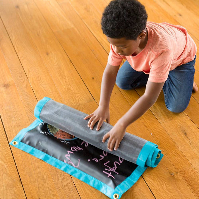 Young child rolling up a portable chalk mat with turquoise edges and corner grommets on a wooden floor, featured in Children's Toys and Indoor Play collections.