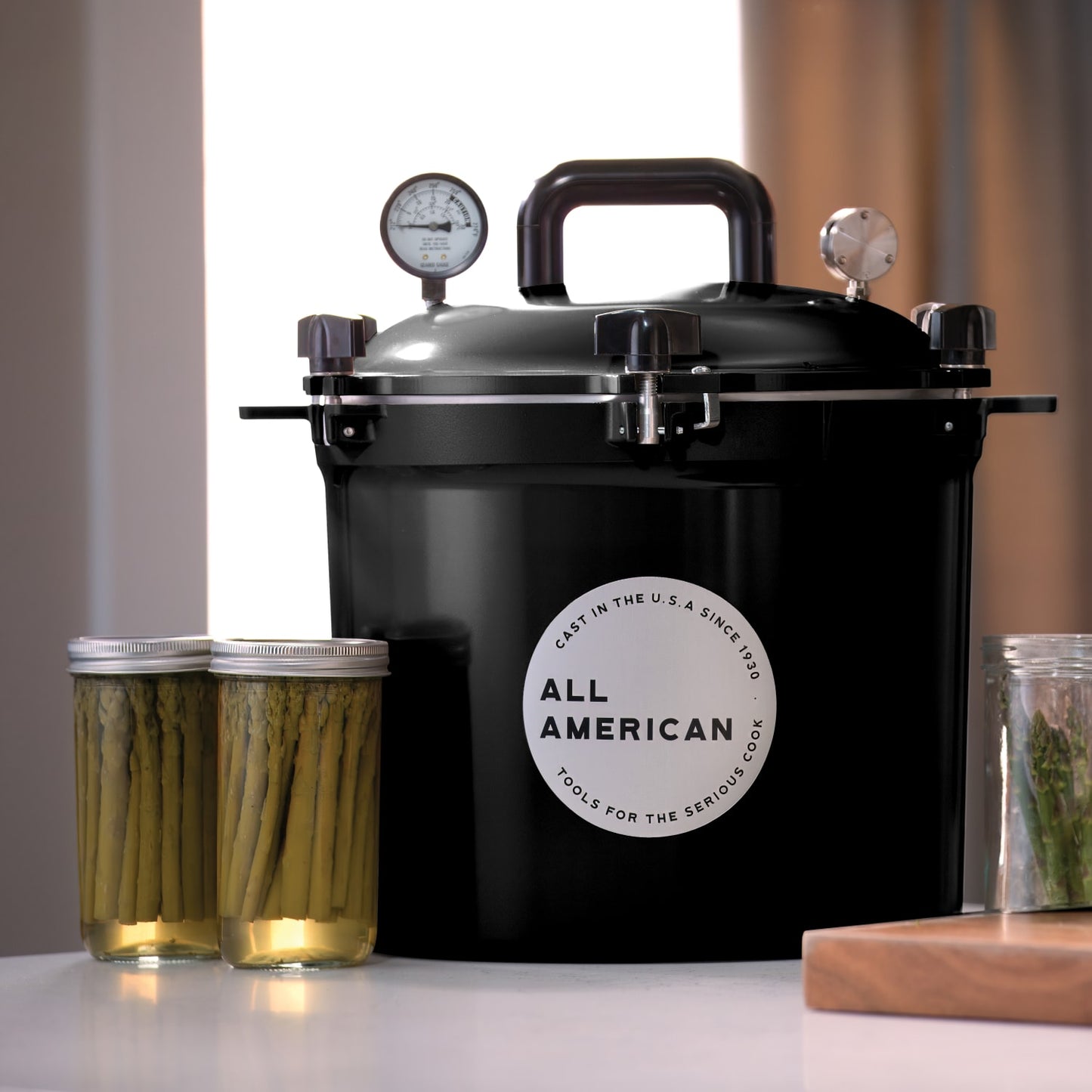 Black pressure canner with secure locking lid and pressure gauges, designed for safe food preservation in Kitchen & Food, Food Preservation, and USA Made collections, accompanied by jars of preserved asparagus.