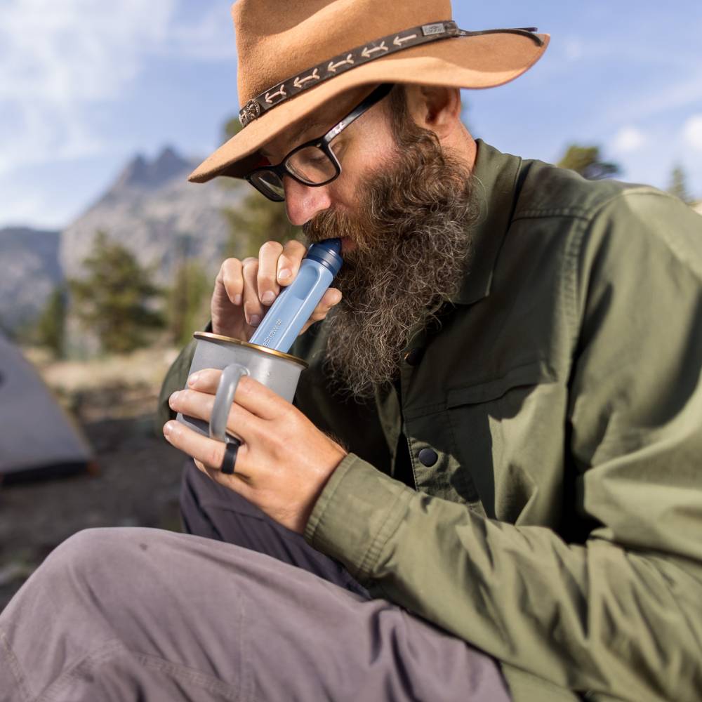 Bearded man wearing glasses and a wide-brimmed hat drinking filtered water through a blue water filter straw from a metal camping cup in an outdoor mountain setting, part of camping and emergency supplies collections.