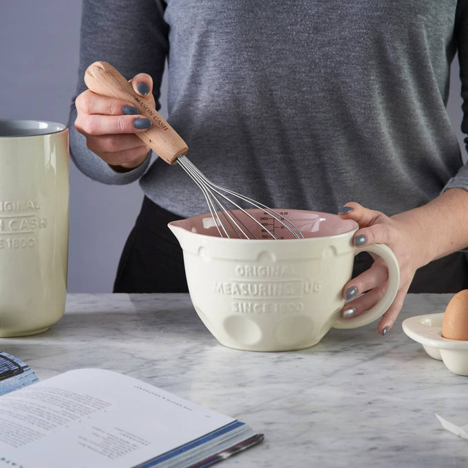 Person using a wooden-handled whisk to mix ingredients in a cream-colored Mason Cash measuring jug with a pink interior on a marble countertop, set among kitchenware in Kitchen & Food, Cookware, Baking Supplies, Food Prep, Measuring, Best Sellers, Promo Eligible collections.