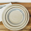 Top view of three handmade stoneware plates with double blue stripes on natural beige ceramic, stacked on a wooden surface with a white cloth nearby, part of Kitchen & Food, Dinnerware, Holiday Hosting, Storage and Serving, USA Made, Best Sellers, and Promo Eligible collections.