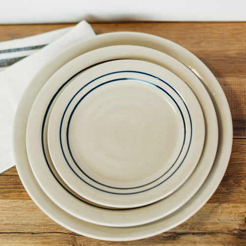 Top view of three handmade stoneware plates with double blue stripes on natural beige ceramic, stacked on a wooden surface with a white cloth nearby, part of Kitchen & Food, Dinnerware, Holiday Hosting, Storage and Serving, USA Made, Best Sellers, and Promo Eligible collections.