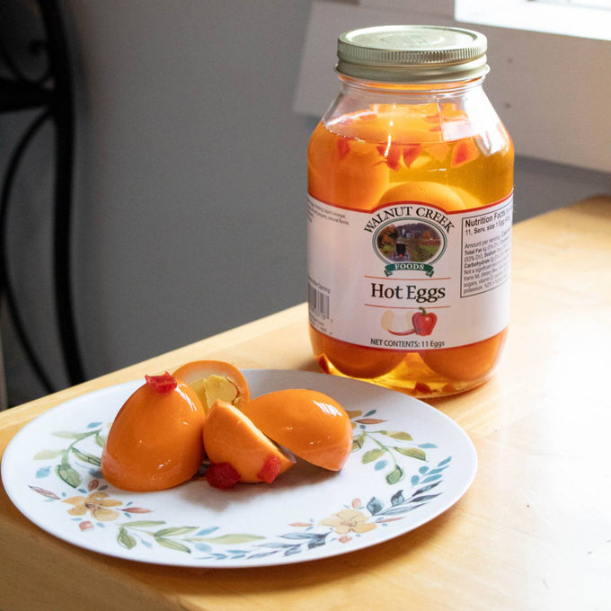 Glass jar of handcrafted hot pickled eggs in spicy brine with visible red pepper pieces, displayed next to peeled eggs on a decorative plate, ideal for Kitchen & Food, Pantry, USA Made, Best Sellers, and Promo Eligible collections.
