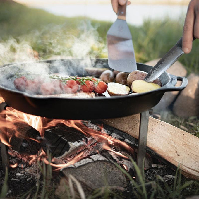 Cast iron fire skillet with two side handles cooking a meal of tomatoes, potatoes, and other vegetables over an open campfire, suitable for farm, kitchen, camping, cookware, and cast iron use.