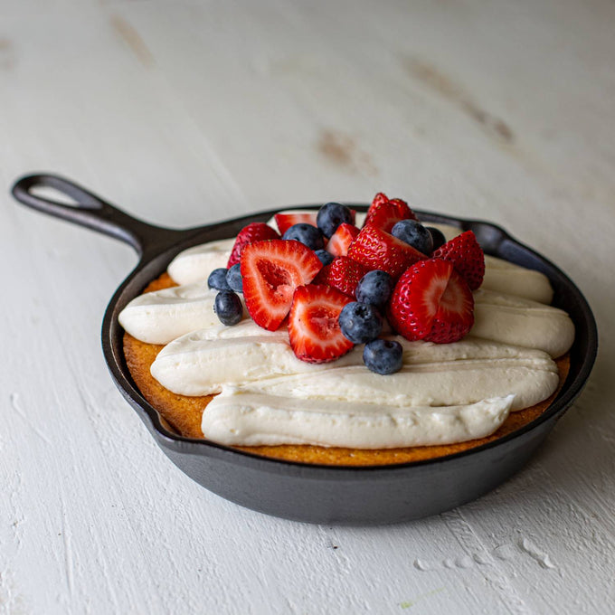 Handcrafted black pre-seasoned cast iron skillet holding a golden baked dessert topped with whipped cream, fresh strawberries, and blueberries for Kitchen & Food, Cookware, Cast Iron, Best Sellers, Promo Eligible collections.