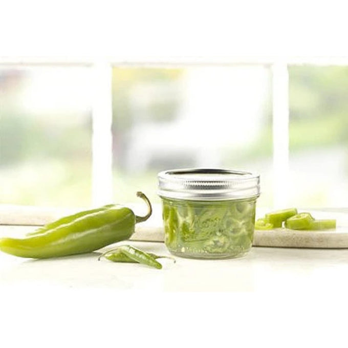Clear glass half-pint wide mouth canning jar with silver lid filled with sliced green chilies, displayed on a light surface with whole and cut chilies nearby, ideal for Kitchen & Food and Food Preservation collections.