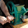 Person using a heavy-duty cast iron grain mill with a green finish and wooden handle on a wooden table, ideal for Kitchen & Food, Baking Supplies, and Grain Mills collections.